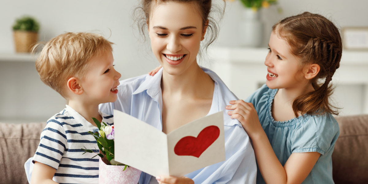 Excited small kids and young woman with flowers reading handmade festive card in light apartment Pleased mother and little children with bouquet of fresh flowers embracing on sofa while reading postcard with red printed heart at home